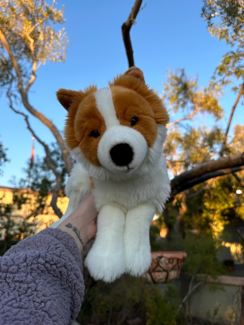 May include: A plush dog toy with a white body, brown ears, and a brown and white face. The toy has a black nose and is held up against a blue sky with tree branches in the background.