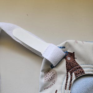 May include: A beige bib featuring a brown leopard print pattern with a white hook-and-loop fastener strap. The bib has a circular neck opening with blue stitching around the edge and a soft, light blue fabric lining on the interior side.