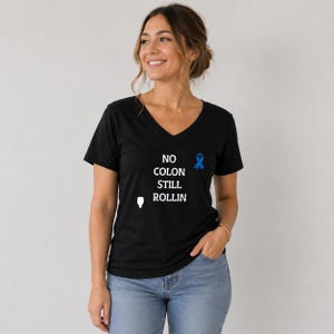 May include: A black V-neck t-shirt featuring the white text NO COLON STILL ROLLIN, a small white icon of a medical bag, and a blue awareness ribbon graphic on the chest. The shirt is paired with light blue denim jeans with a visible metal button.