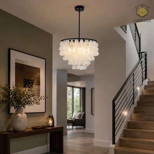 May include: A modern chandelier with a black frame and a cascade of white, shell-like elements. The light fixture hangs in a hallway with a staircase and artwork. The brand name "LUNESSA" is visible.