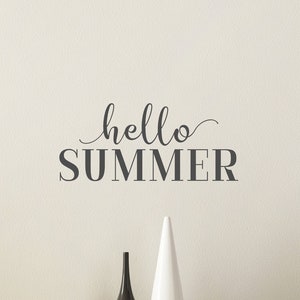 May include: A white wall with a gray vinyl decal that says "hello summer" in cursive lettering. The decal is above a wooden dresser with two white doors and silver handles.