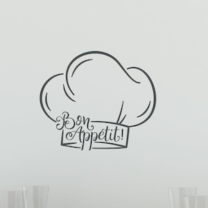 May include: A dark gray wall decal featuring a chef hat outline with the text Bon Appétit! written in a cursive script across the base. The decal is displayed on a plain wall above a wooden table set with white bowls and clear glass stemware.
