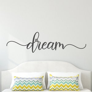 May include: A white bed with a yellow bedspread and two pillows with a chevron pattern. The headboard is white and the wall behind the bed is gray. The word "dream" is written in black cursive on the wall above the bed.