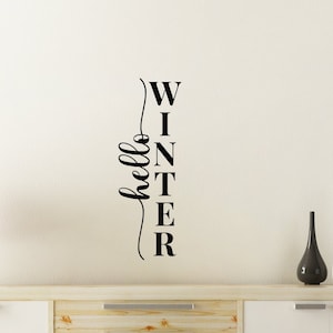 May include: A white wall with a black vinyl decal that reads "hello winter" in a vertical script font. The decal is on a wall above a wooden dresser with white doors.