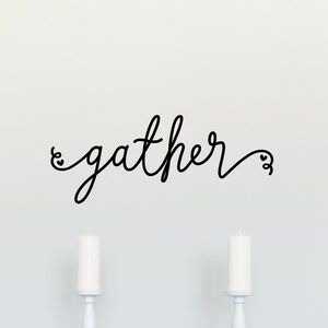 May include: A white dining room table with four white chairs and a teal blue tablecloth. The wall behind the table has a black vinyl decal that reads "gather" in a cursive font.