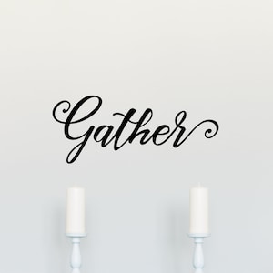 May include: A dining room scene featuring a white table and chairs, set against a white wall. The word "Gather" is written in elegant black script above the table. Two white candles sit on the table.