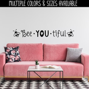 May include: A pink sofa with two pillows, a coffee table with a book and a small plant, and a black wall decal that reads "Bee You Tiful" with a bee on each side of the text.