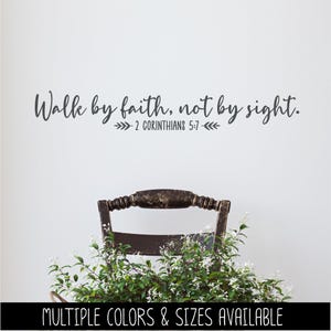 Pode incluir: Um adesivo de parede cinza-escuro com o texto em cursivo e impresso Walk by faith, not by sight. 2 CORINTHIANS 5:7 e pequenos ícones decorativos de folhas em ambos os lados da referência bíblica, posicionado acima de uma cadeira de madeira.