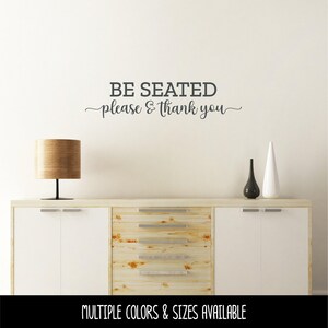 May include: A dark gray wall decal featuring the text BE SEATED in a bold, sans-serif font, followed by the text please & thank you in a cursive script font. The decal is applied to a plain wall above a wooden sideboard with a lamp and vases.