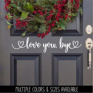 May include: A white vinyl decal applied to a dark gray door featuring the cursive text "love you, bye" flanked by two heart shapes. Above the decal, a decorative wreath with green leaves, pinecones, and red berries hangs on the door surface.