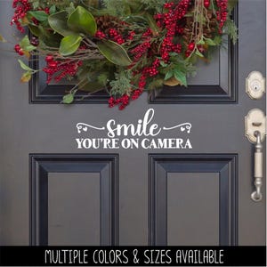 May include: A white vinyl decal applied to a dark gray door featuring the text "smile" in a cursive script above the text "YOU'RE ON CAMERA" in a bold sans-serif font, positioned below a festive holiday wreath with red berries and green leaves.