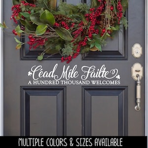 May include: A white vinyl decal applied to a dark gray door featuring the Irish text Céad Míle Fáilte and the English translation A HUNDRED THOUSAND WELCOMES in a decorative script font, accented by small shamrock icons on each side.
