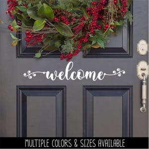 May include: A white vinyl decal featuring the word welcome in a cursive script font with decorative swirls on each side. The decal is applied to a dark gray door positioned below a festive wreath made of green leaves, pinecones, and red berries.