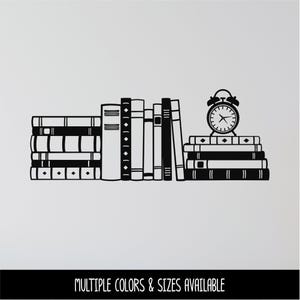 May include: A black wall decal featuring a stack of books arranged horizontally and vertically, topped with an analog alarm clock. The design includes various book spine details and the text "MULTIPLE COLORS & SIZES AVAILABLE" at the bottom.