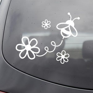 May include: White vinyl decal of a bee with a string of beads leading to three white flowers. The bee has a striped body and is flying.