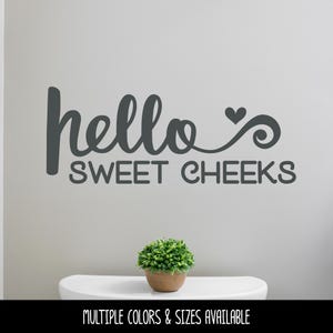 May include: A dark gray wall decal featuring the text "hello" in a cursive script followed by a small heart and a swirl, with the words "SWEET CHEEKS" in a bold, rounded sans-serif font positioned directly underneath the first line of text.