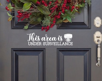 May include: A white vinyl decal applied to a dark gray door featuring the text This area is UNDER SURVEILLANCE with a security camera icon. The door has a silver handle and lock, with a festive holiday wreath of pine, berries, and cones above.
