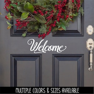 May include: A dark gray door featuring a white vinyl decal with the word Welcome in a cursive script font. Above the text, a holiday wreath displays green magnolia leaves, brown pinecones, and clusters of bright red berries on thin branches.