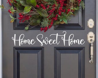 May include: A dark gray door featuring a white vinyl decal with the cursive text Home Sweet Home. Above the text, a decorative wreath is displayed, composed of green magnolia leaves, pine cones, and clusters of bright red berries.