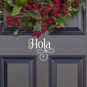 May include: A dark gray front door featuring a white vinyl decal with the word Hola in a cursive script font. A festive wreath with green leaves, red berries, and pinecones hangs above the text. The door has a brass handle and deadbolt lock.