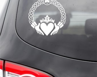 Puede incluir: Un adhesivo de vinilo blanco con un diseño de Claddagh que muestra un corazón, una corona y dos manos, encerrado en un borde circular de nudo celta rematado con un trébol. El adhesivo está colocado en la luneta trasera de un vehículo oscuro.