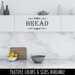 May include: A white kitchen cabinet door featuring a dark gray vinyl decal with the word BREAD in a serif font, framed by decorative scrollwork flourishes above and below the text. The cabinet is positioned above a marble countertop and backsplash.