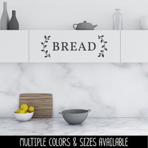 May include: A dark gray vinyl wall decal featuring the word BREAD in capital letters, flanked by two vertical leafy vine illustrations. The decal is applied to a white kitchen cabinet door, positioned above a marble countertop and backsplash.