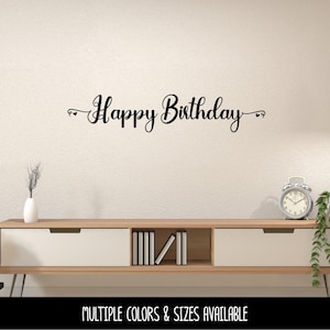 May include: A black wall decal featuring the text Happy Birthday in a cursive script font with small heart accents on both ends. The lettering is displayed horizontally on a plain wall surface above a wooden media console unit.