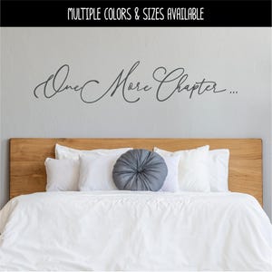 Pode incluir: Um adesivo de parede cinza-escuro com o texto em cursivo "One More Chapter..." aplicado em uma parede cinza-claro acima de uma cabeceira de madeira. O texto está escrito em uma fonte fluida e elegante, centralizada horizontalmente na parede.