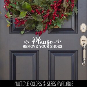 May include: A white vinyl decal applied to a dark gray door featuring the text Please REMOVE YOUR SHOES in a combination of script and block lettering. Above the decal, a festive holiday wreath with green leaves, pinecones, and red berries.
