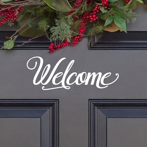 May include: A white vinyl decal featuring the word Welcome in a cursive script font applied to a dark gray paneled door. Above the text, a portion of a decorative wreath is visible, showcasing green leaves and clusters of bright red berries.