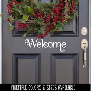 May include: A dark gray door featuring a white vinyl decal with the word Welcome in a stylized script font. Above the text, a decorative wreath is displayed, composed of green magnolia leaves, pine cones, and clusters of bright red berries.