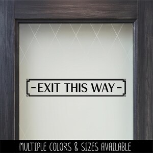 May include: A black rectangular vinyl decal applied to a glass door with a frosted finish. The decal features a decorative border with geometric corners and the text "EXIT THIS WAY" centered between two horizontal dashes in a serif font.