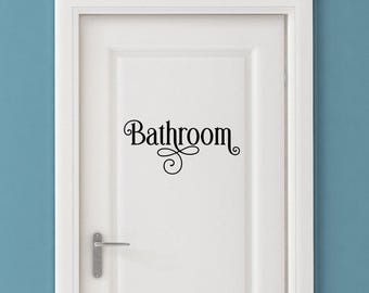 May include: A white interior door featuring a black vinyl decal with the word Bathroom written in a stylized, cursive-inspired font with decorative flourishes underneath the text. The door includes a silver metal handle on the left side.