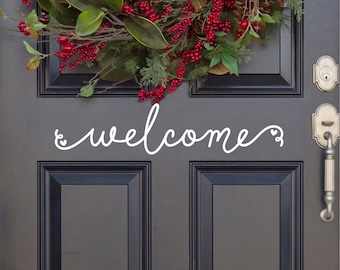 May include: A white vinyl decal with the word welcome in a cursive script font, featuring small heart accents at the beginning and end. The decal is applied to a dark gray door positioned below a festive wreath with green leaves and red berries.