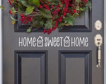 May include: A dark grey door featuring a white vinyl decal that reads HOME SWEET HOME with two house icons containing hearts. Above the text, a wreath made of green magnolia leaves, pine cones, and clusters of bright red berries is displayed.