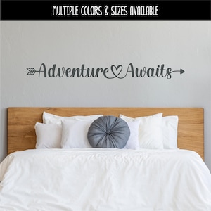 May include: A gray wall decal with the words "Adventure Awaits" in a script font, with an arrow and heart design. The decal is above a wooden headboard and white bedding. The text "MULTIPLE COLORS & SIZES AVAILABLE" is at the top.