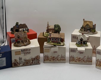 Może przedstawiać: Osiem miniaturowych domków w różnych stylach, każdy na małej podstawie z pudełkiem. Pudełka są wszystkie białe z nadrukiem "Lilliput Lane". Domki są w różnych kolorach i mają różne detale, takie jak kominki, okna i drzwi.