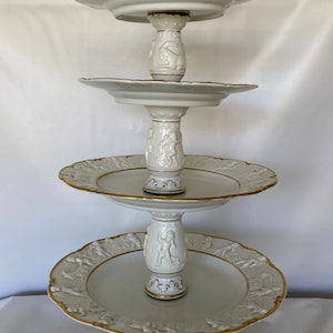Puede incluir: Un soporte de servicio de porcelana blanca de cuatro niveles con detalles dorados. Cada nivel presenta un diseño en relieve de querubines y motivos florales. El nivel superior está adornado con una figura de querubín que sostiene una corona.