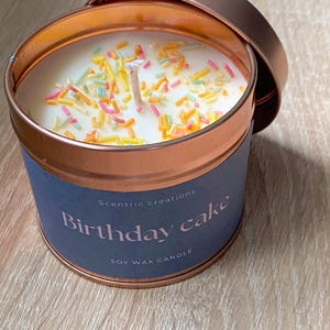 以下が含まれることがあります： 銅製の缶に入った大豆ワックスのキャンドル。白いキャンドルにカラフルなスプリンクルがトッピングされています。ラベルには「Scentric creations Birthday cake Soy Wax Candle」と書いてあります。