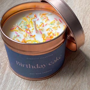 以下が含まれることがあります： 銅色のキャンドル缶に、青いラベルが貼ってあり、「Scentric creations Birthday cake」と「Soy Wax Candle」と書かれています。キャンドルは白く、カラフルなスプリンクルがトッピングされています。