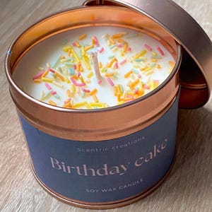 以下が含まれることがあります： 銅色のキャンドル缶に、青いラベルが貼ってあり、「Scentric creations Birthday cake SOY WAX CANDLE」と書いてあります。キャンドルは白で、カラフルなスプリンクルがかけられています。