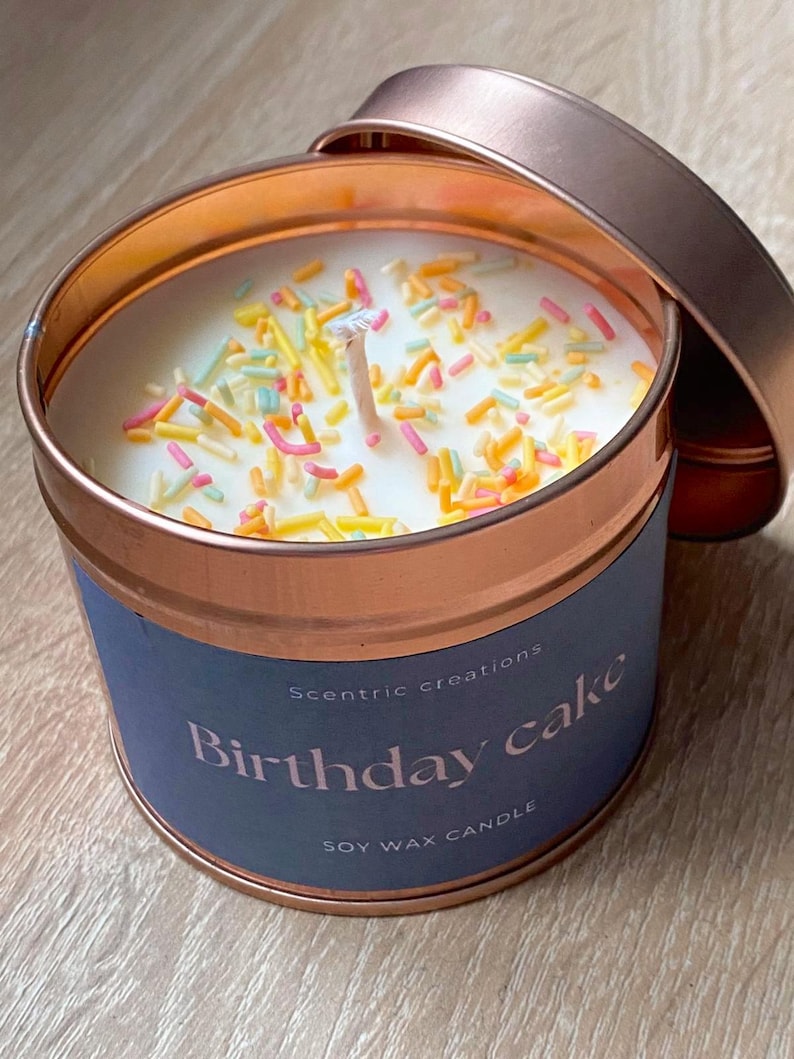 以下が含まれることがあります： 銅色のキャンドル缶に、青いラベルが貼ってあり、「Scentric creations Birthday cake SOY WAX CANDLE」と書いてあります。キャンドルは白で、カラフルなスプリンクルがかけられています。
