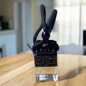 May include: A small, square glass bottle containing a clear liquid, topped with a dark navy blue, perforated plastic cap. A matching dark navy blue braided cord is looped through the top of the cap, secured by a small, round navy blue bead.