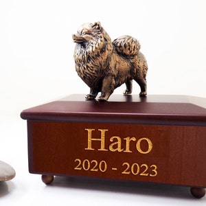 May include: A wooden pet urn with a gold-coloured Pomeranian figurine on top. The urn has a dark wood finish and is engraved with the name "Haro" and the dates "2020 - 2023".