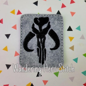 May include: A grey felt patch featuring a black Mandalorian skull emblem. The patch is rectangular with rounded corners and white stitching. The text "Wandering Hero Stitch" is at the bottom.