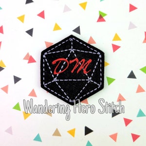 Może przedstawiać: Czarna filcowa naszywka w kształcie kostki D20, z białymi przeszyciami i literami "DM" wyhaftowanymi na czerwono. Naszywka znajduje się na białym tle z kolorowymi trójkątami. Pod naszywką znajduje się napis "Wandering Hero Stitch".