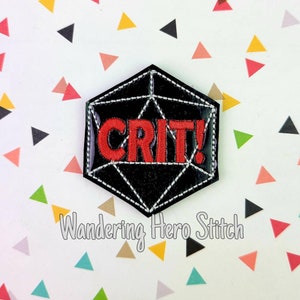 May include: A black, geometric patch featuring the word "CRIT!" in red, stitched lettering. The patch is shaped like a twenty-sided die, with white stitching outlining the edges and interior lines. The background is white with colourful triangle shapes.