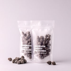May include: Two clear, resealable stand-up pouches filled with small, brown, candied pine cones. The white text on the front of the bags reads: HANDMADE CANDIED PINE CONES, take a bite of the forest. Several loose pine cones are scattered in front.