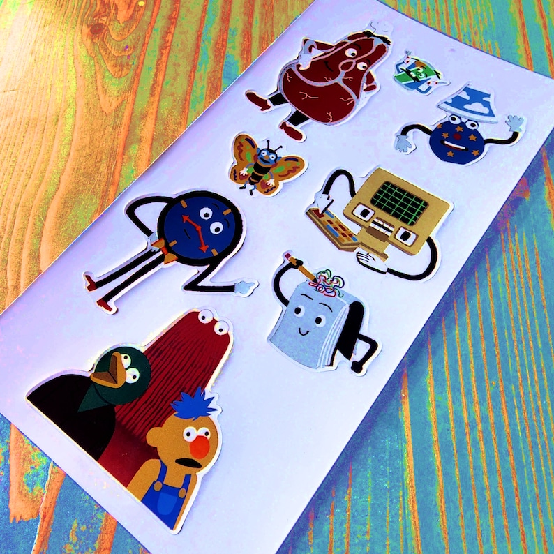 K&ouml;nnte beinhalten: Ein Set mit bunten Comic-Charakter-Stickern mit einer Uhr, einem Schmetterling, einem Buch, einem Computer, einem Fleischlaib und einem Vogel. Die Sticker sind auf wei&szlig;em Hintergrund.
